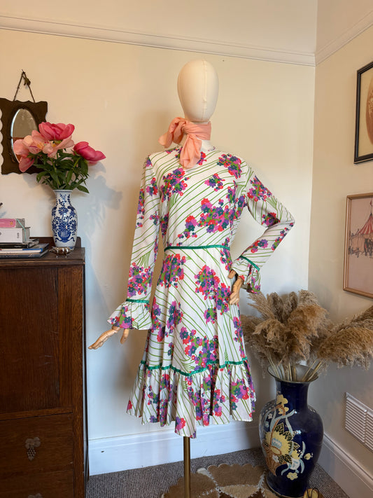 
Vintage 1970s white floral midi dress with bright pink, purple and green abstract flower print, long sleeves with flared cuffs, a fitted waist and ruffled hem displayed on a mannequin with a peach neck scarf.
